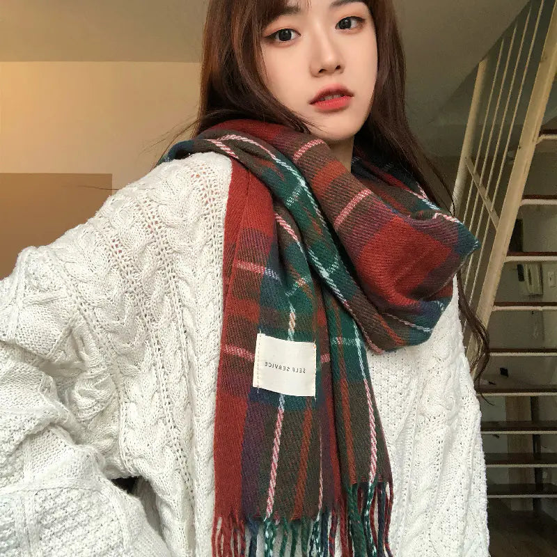 A woman wearing a soft plaid scarf with tassels in red, green, and dark tones over a chunky white knit sweater.