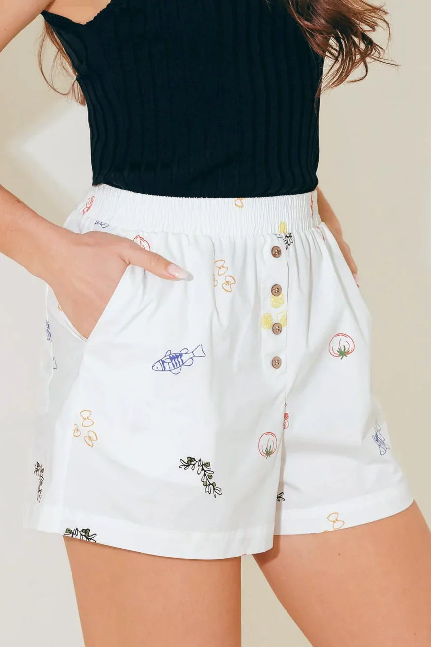Close-up view of Flying Tomato white woven shorts with multi-color food and nature graphic print, showing smocked waistband, front buttons, and side pockets.