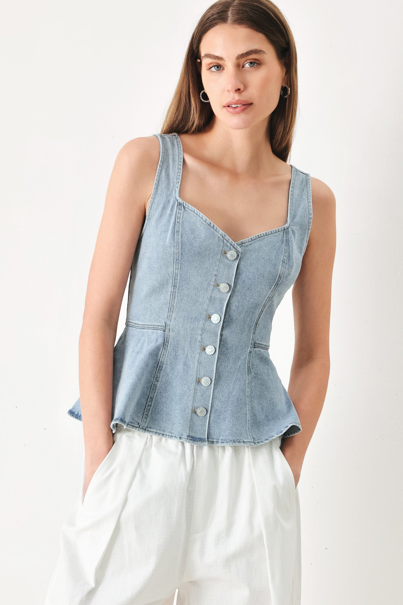 A woman wearing a light blue denim sleeveless peplum top with a button-down front and Queen Anne neckline.