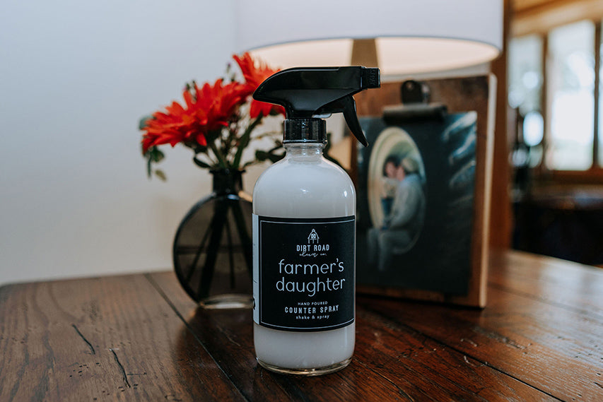 A bottle of Farmer's Daughter counter spray sitting on a wooden table next to a vase of red flowers.