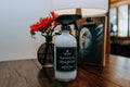 A bottle of Farmer's Daughter counter spray sitting on a wooden table next to a vase of red flowers.