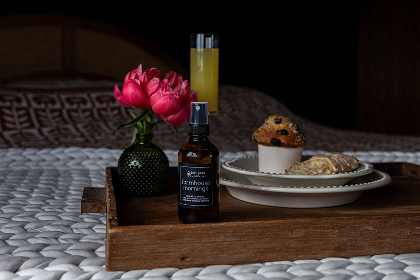 Farmhouse Mornings room spray bottle placed on a rustic wooden tray with a flower vase and pastries.