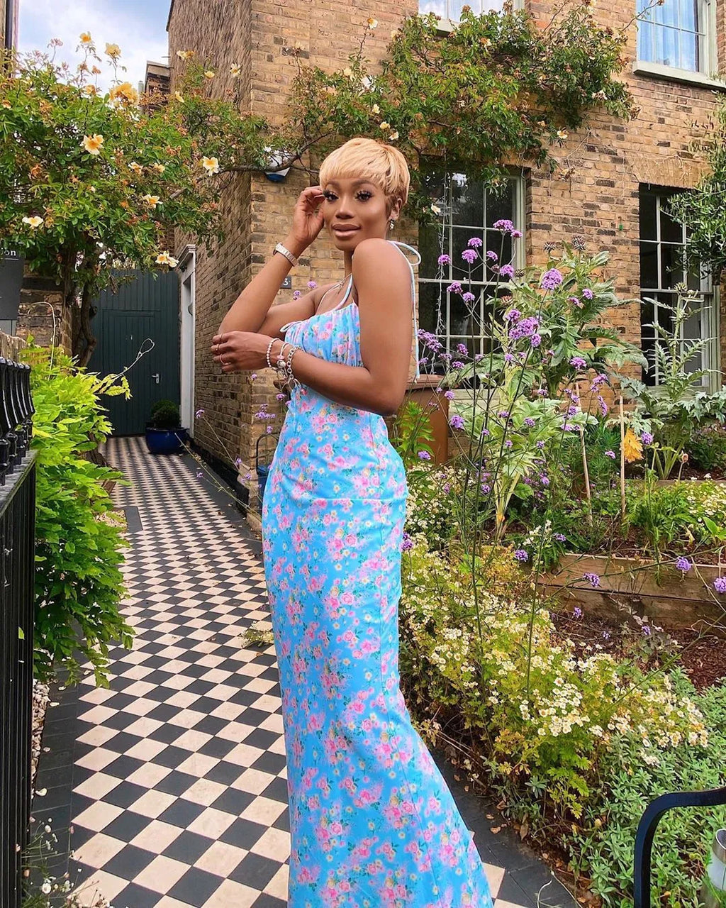 A woman wearing a light blue floral maxi dress with pleated bodice and tie straps standing in a garden setting.