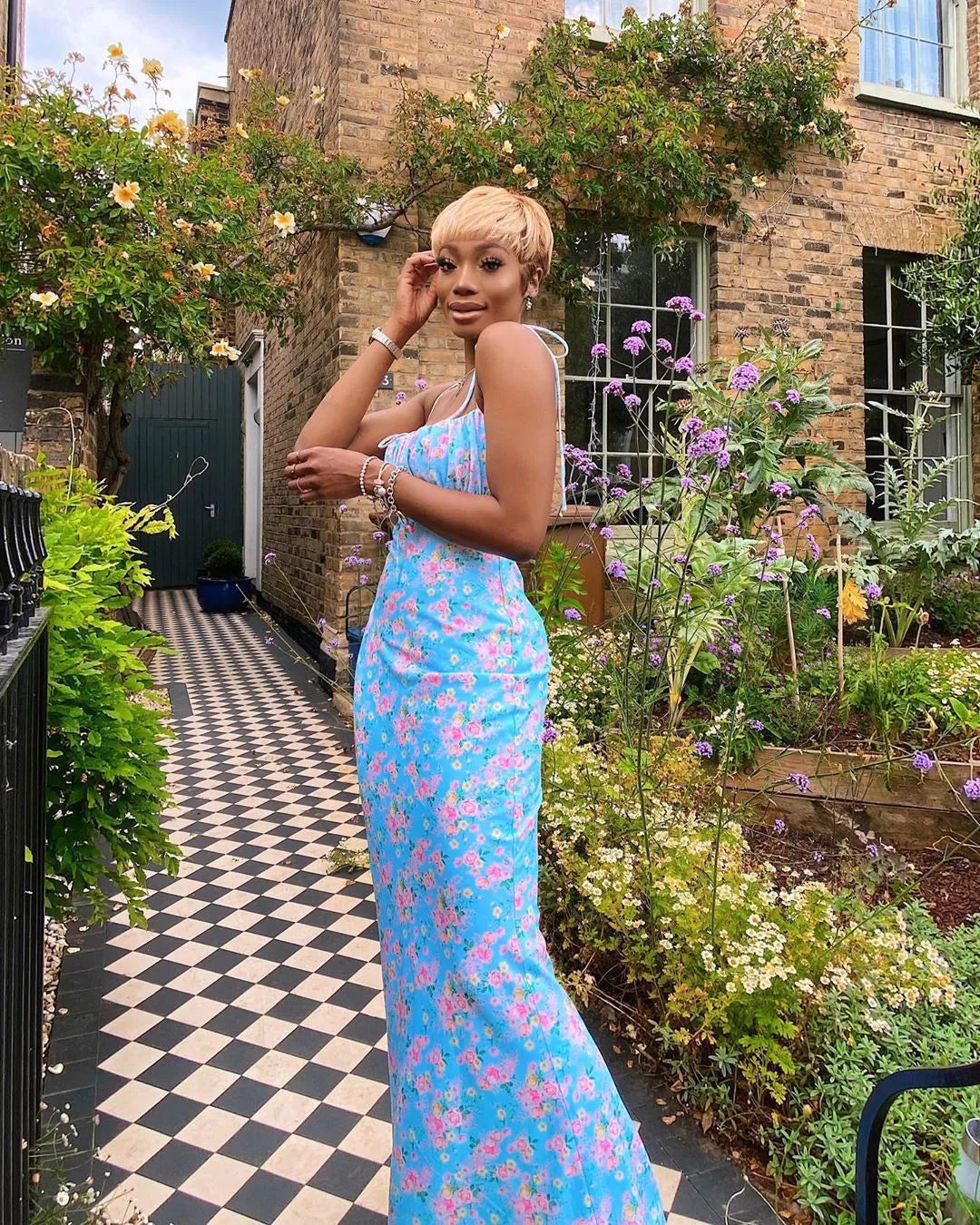 A woman wearing a light blue floral maxi dress with pleated bodice and tie straps standing in a garden setting.