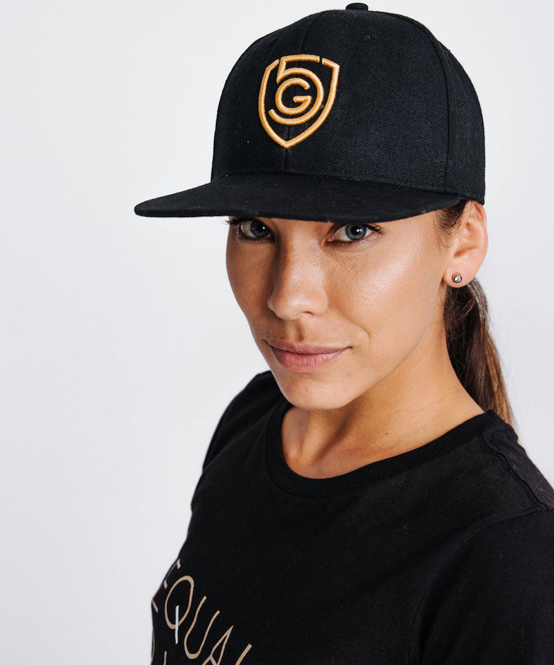 A person wearing a black flat-billed snapback hat with a gold embroidered logo.