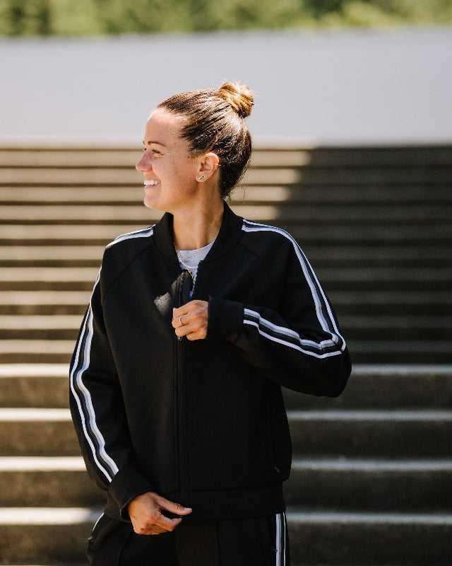 Woman wearing a black Powerstripe track jacket with white side stripes and ribbed cuffs.