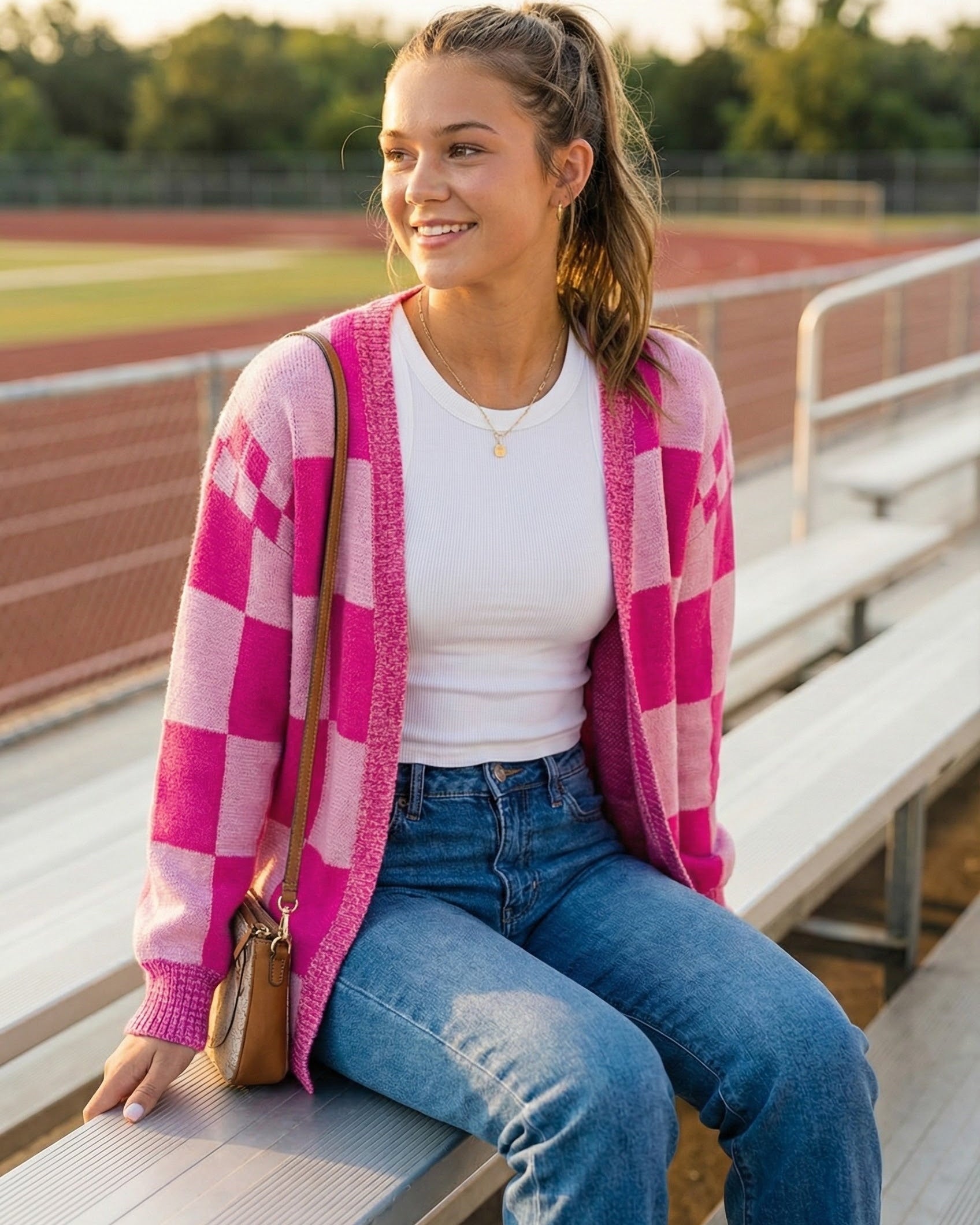 Woman sitting on bleachers wearing a pink checkered cardigan, white shirt, and blue jeans.