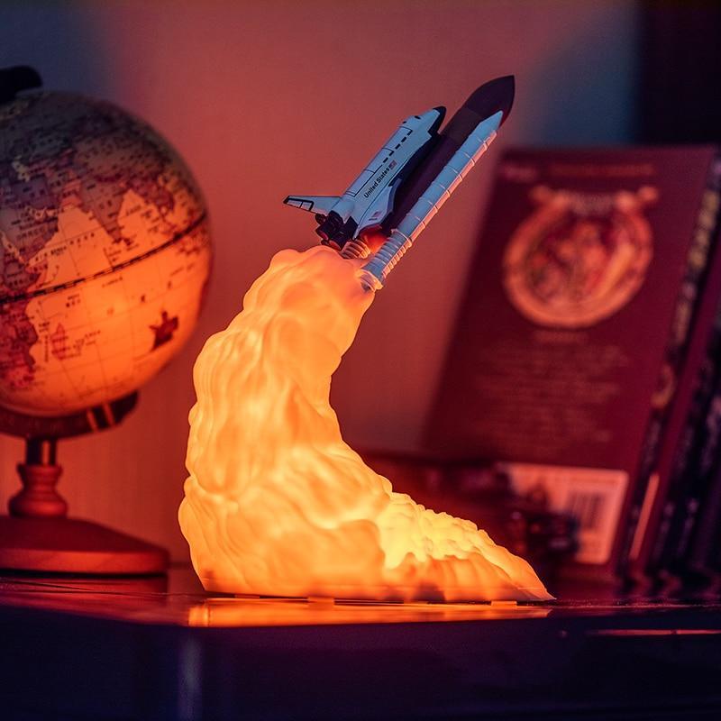 A 3D-printed rocket lamp with white and orange details glowing in a darkened room.