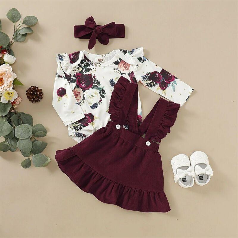 Baby girl wearing a creamy white floral romper jumpsuit with a maroon skirt and matching floral headband.