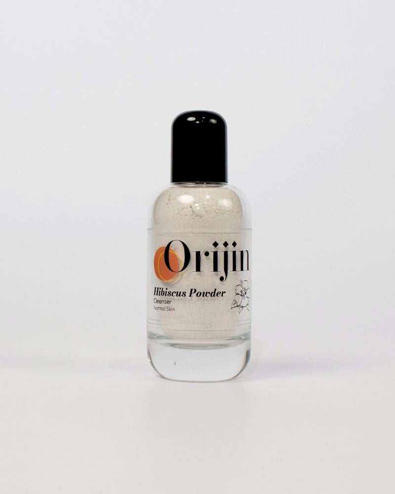 Close-up of Orijin Hibiscus Powder exfoliating cleanser being applied as a dark pink paste to a woman's cheek.