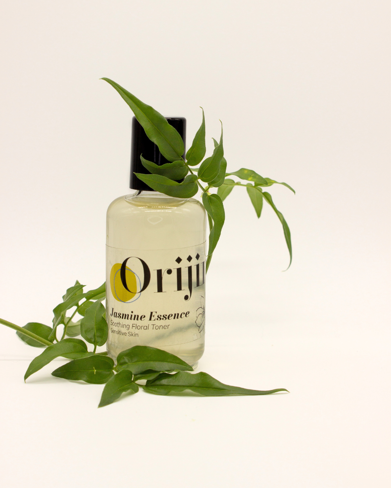 A clear glass bottle of Orijin Jasmine Essence facial toner surrounded by fresh green jasmine leaves.