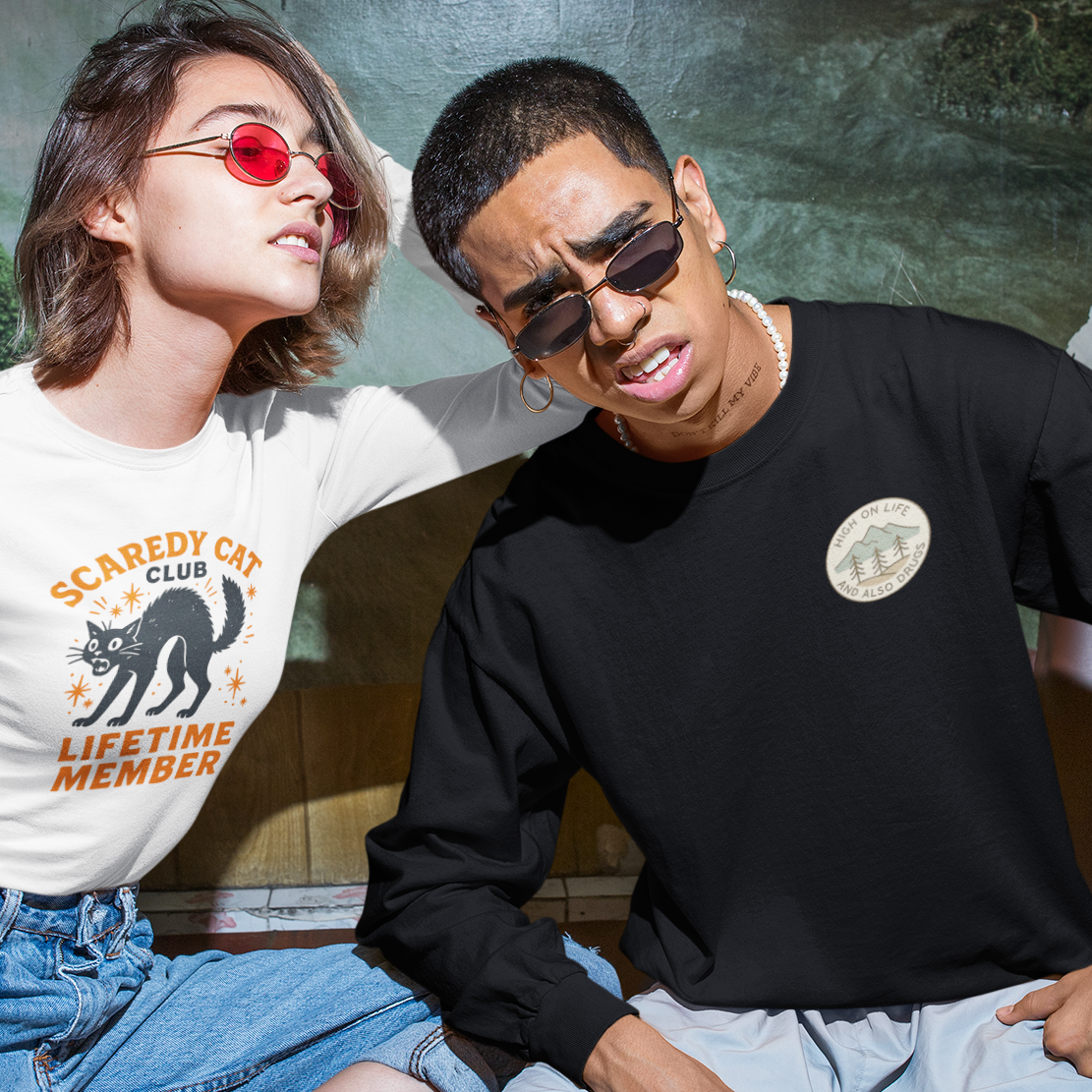 Two models posing together in a styled setting. The model on the left wears a white graphic t-shirt with a black cat illustration and orange text reading 'SCAREDY CAT CLUB LIFETIME MEMBER', paired with blue denim shorts and black boots.