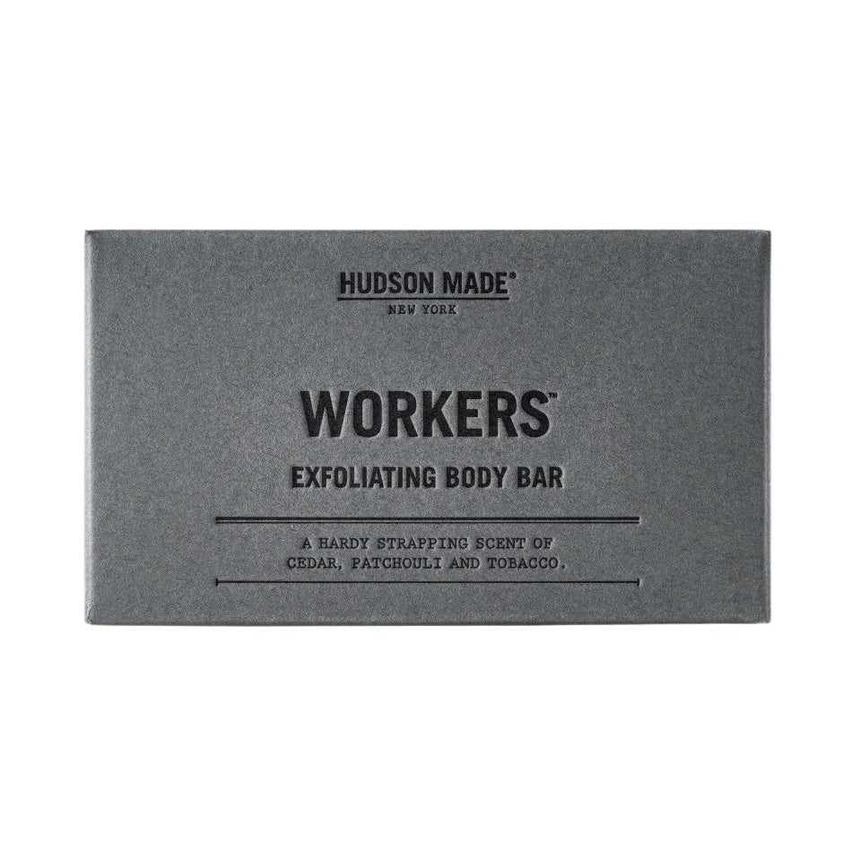 A grey box of Hudson Made Workers Exfoliating Body Bar Soap next to the dark brown, textured soap bar.