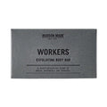 A grey box of Hudson Made Workers Exfoliating Body Bar Soap next to the dark brown, textured soap bar.