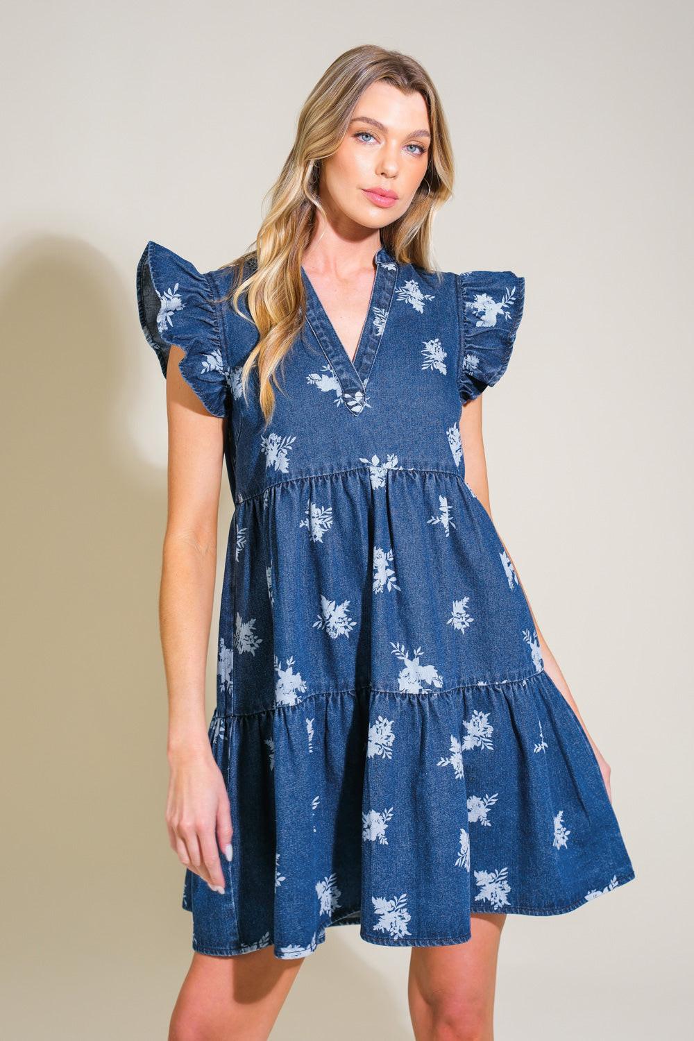 Model wearing a dark wash denim mini dress with a white floral print, featuring ruffled sleeves and a tiered skirt.