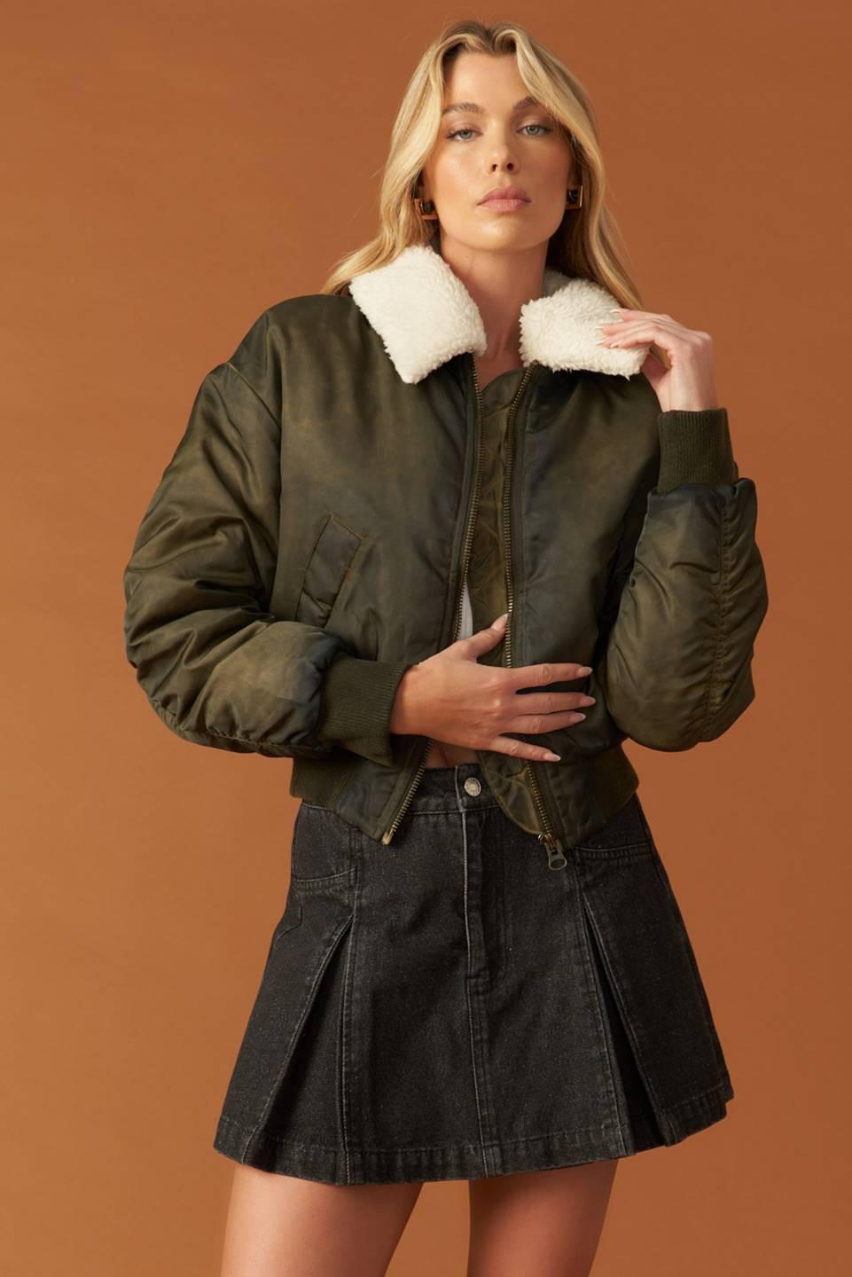 A woman wearing an olive green woven bomber jacket with a white faux shearling collar and zip-up closure.