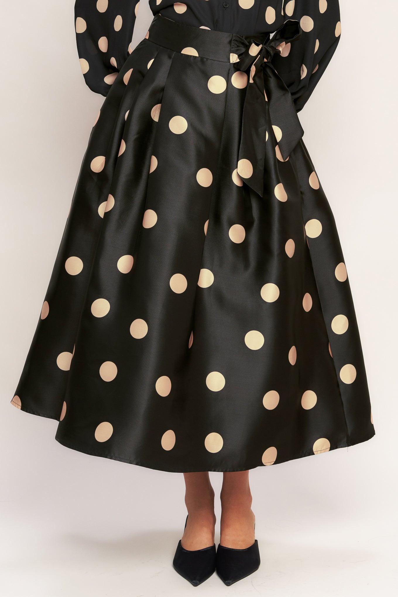 Model wearing a black woven midi skirt with a large cream-colored polka dot pattern, featuring a tie-waist detail and side zipper.