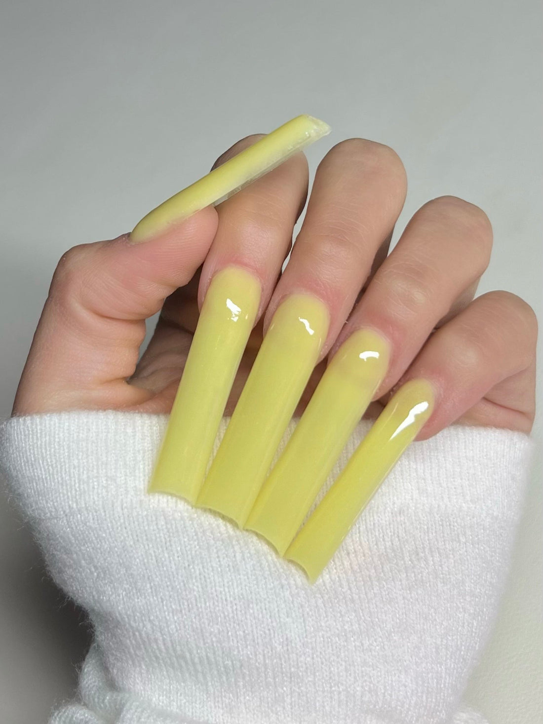 Close up of a hand with long, square-shaped yellow acrylic nails, alongside a container of 2Cute Nails Yellow Daze acrylic powder.