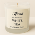 A 10 oz Affinati White Tea signature candle in a frosted glass jar on a light wooden surface next to loose tea leaves.