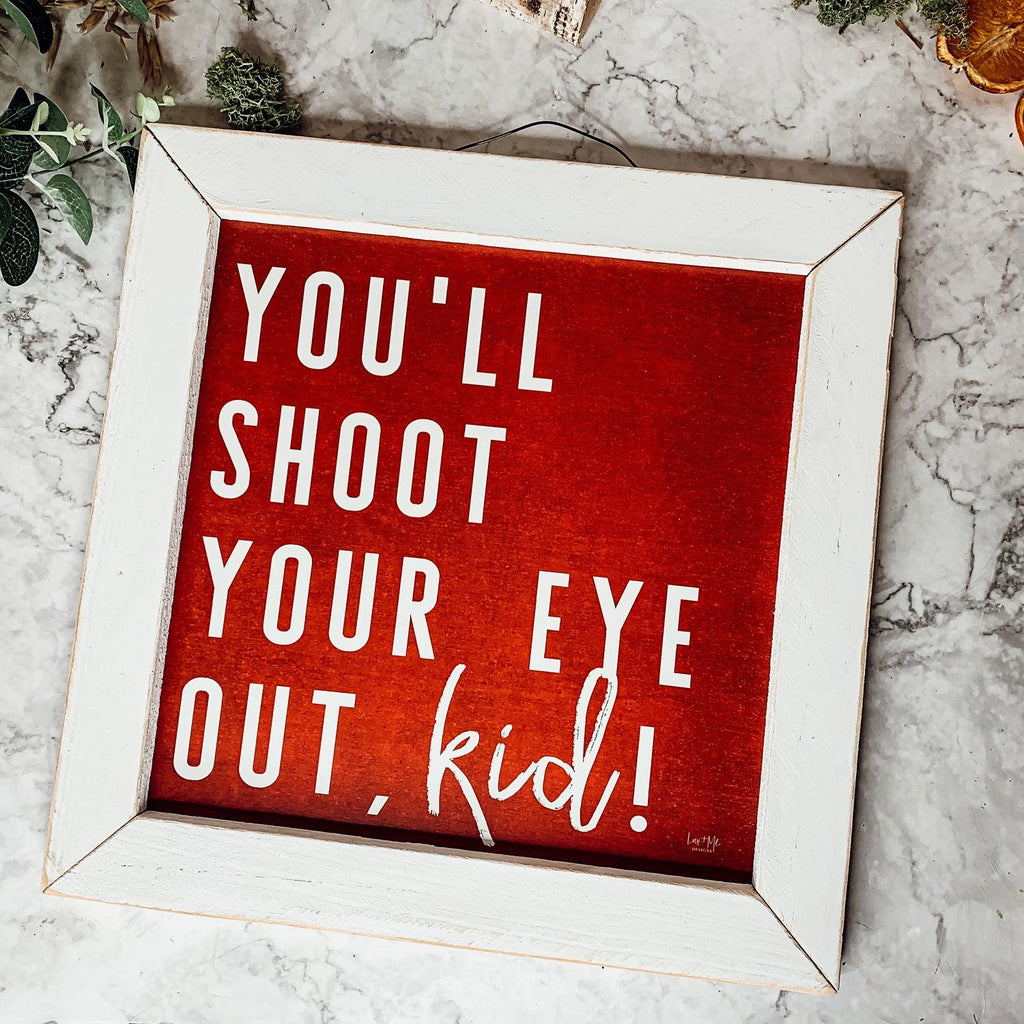 Framed holiday sign with red background and white text reading You Will Shoot Your Eye Out kid in a rustic white frame