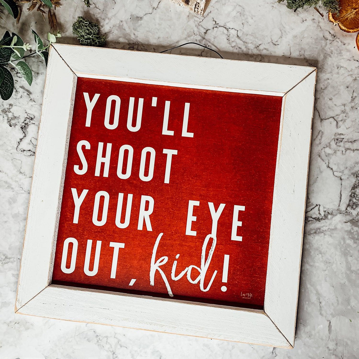 Framed holiday sign with red background and white text reading You Will Shoot Your Eye Out kid in a rustic white frame