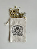 A reusable beige cotton muslin bath soak bag with double drawstring closure, filled with dried chamomile flowers and bath salts.