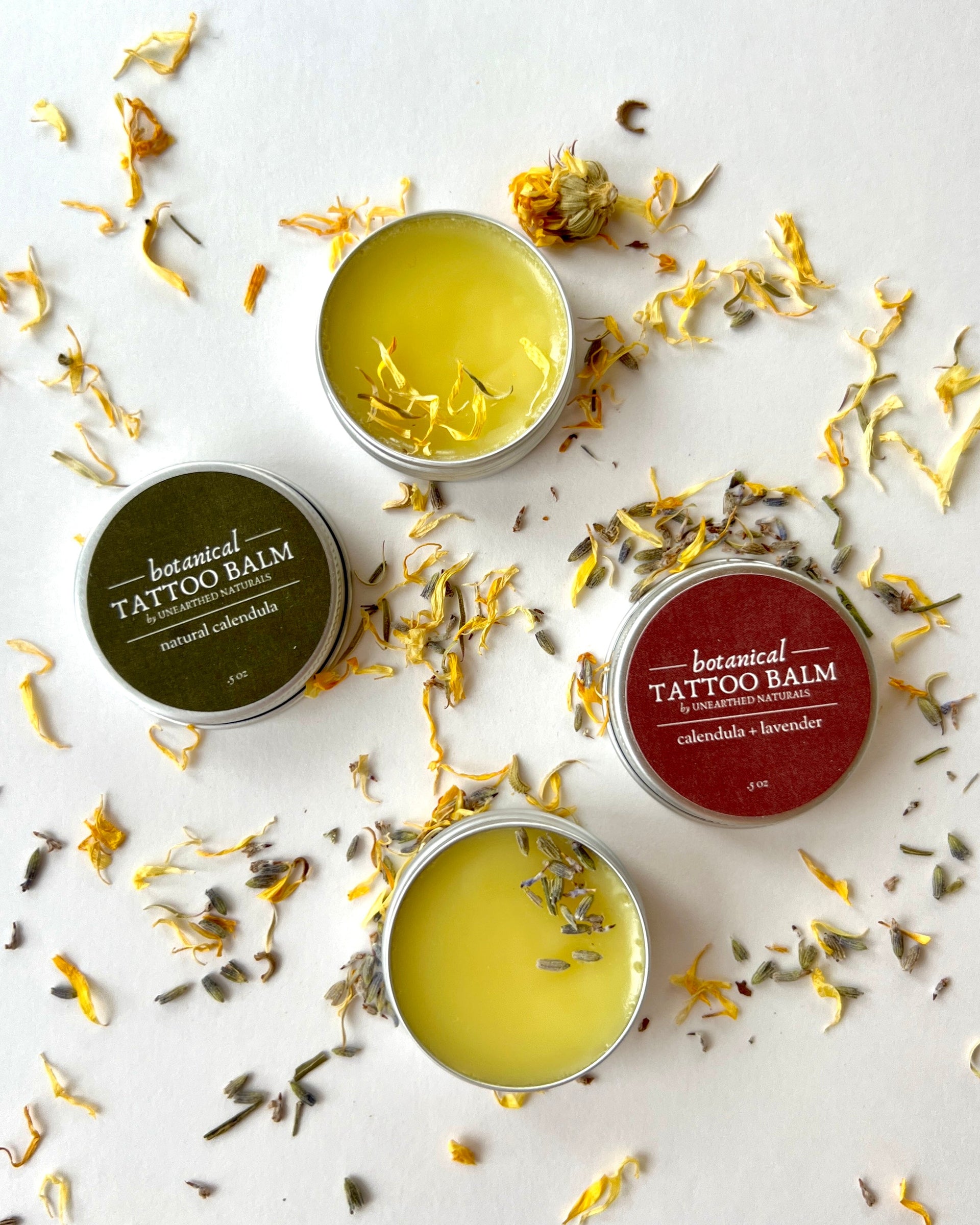 Top view of two round metal tins of botanical tattoo balm surrounded by loose calendula petals and lavender buds.