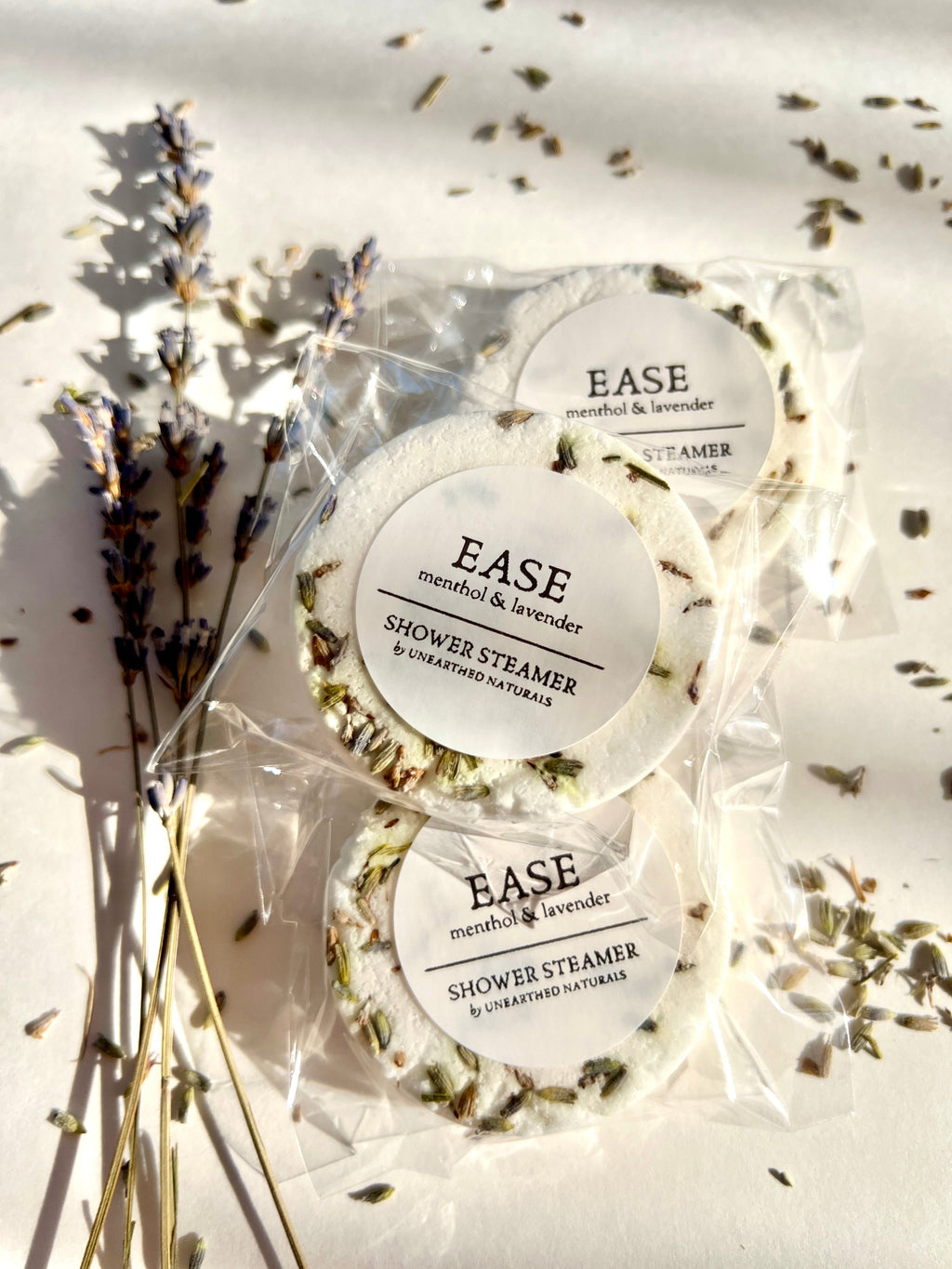 Three circular white lavender and menthol shower steamers wrapped in plastic on a white background with dried lavender sprigs.