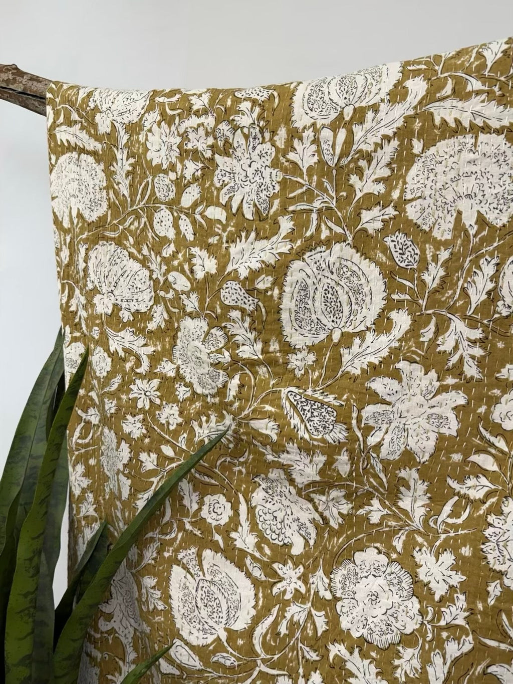 Yellow cotton kantha quilt featuring detailed white floral block print patterns draped over a wooden stand.
