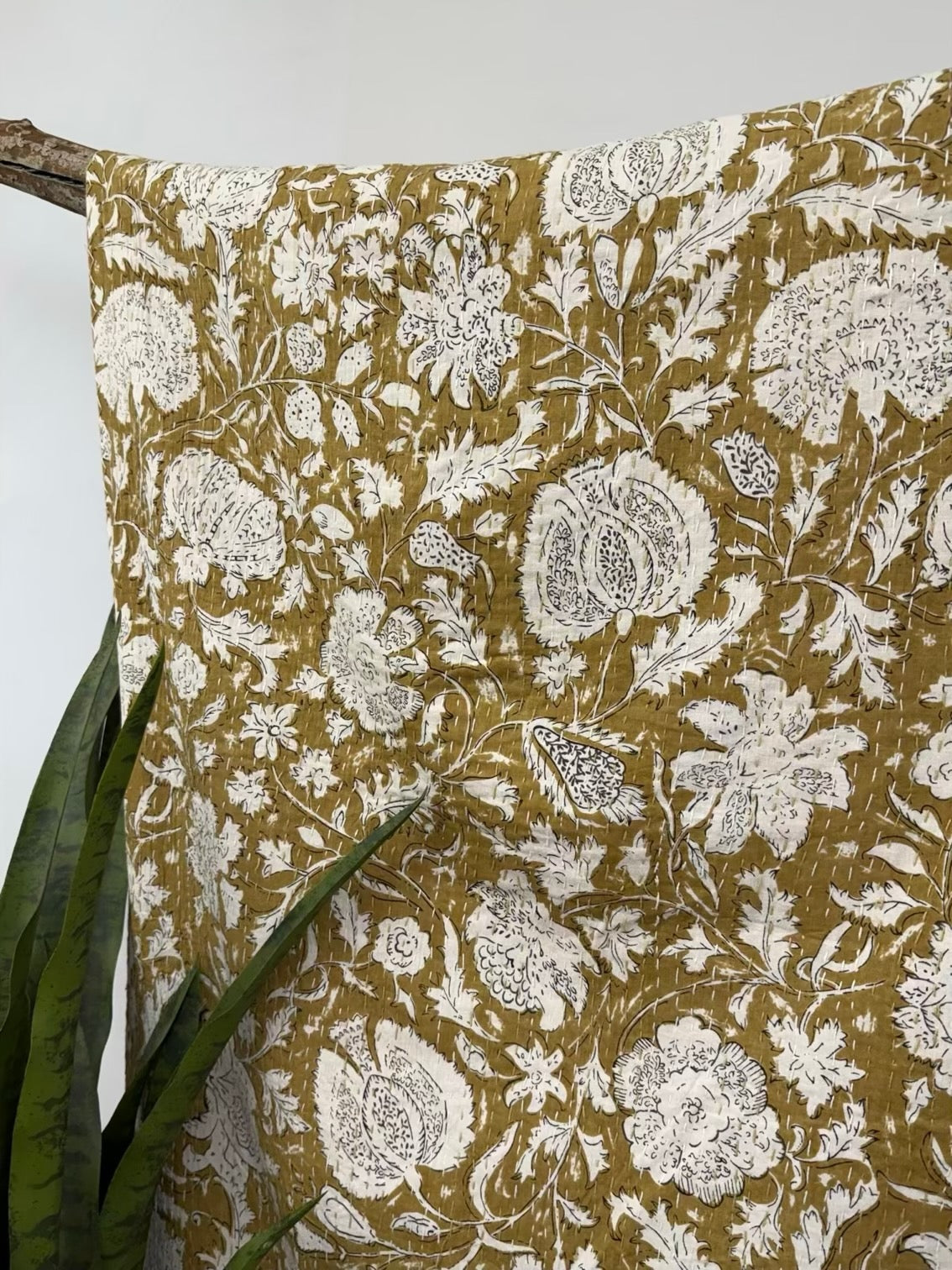 Yellow cotton kantha quilt featuring detailed white floral block print patterns draped over a wooden stand.