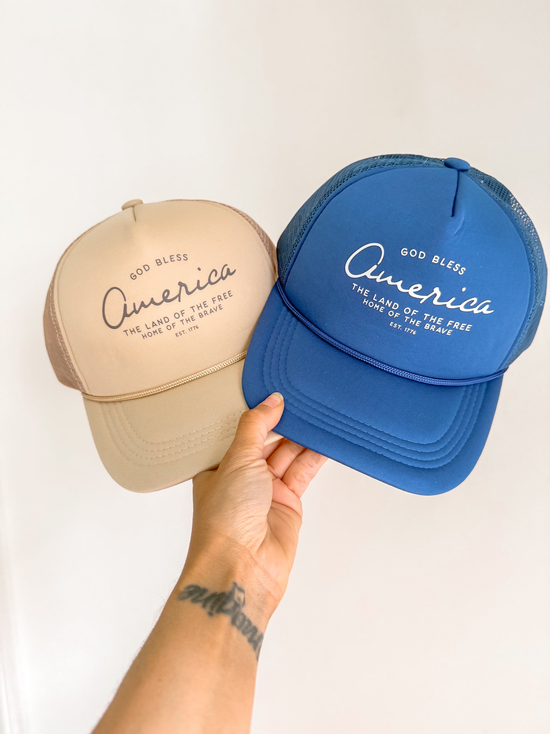 God Bless America trucker hat in black, blue, and beige with foam front panel and adjustable snapback closure.