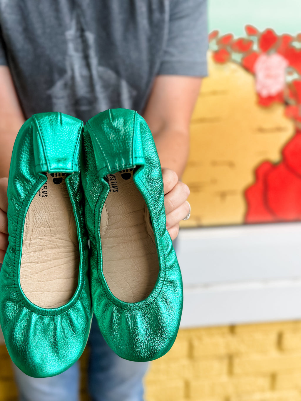 A pair of emerald green metallic foil ballet flats held up against a colorful backdrop.