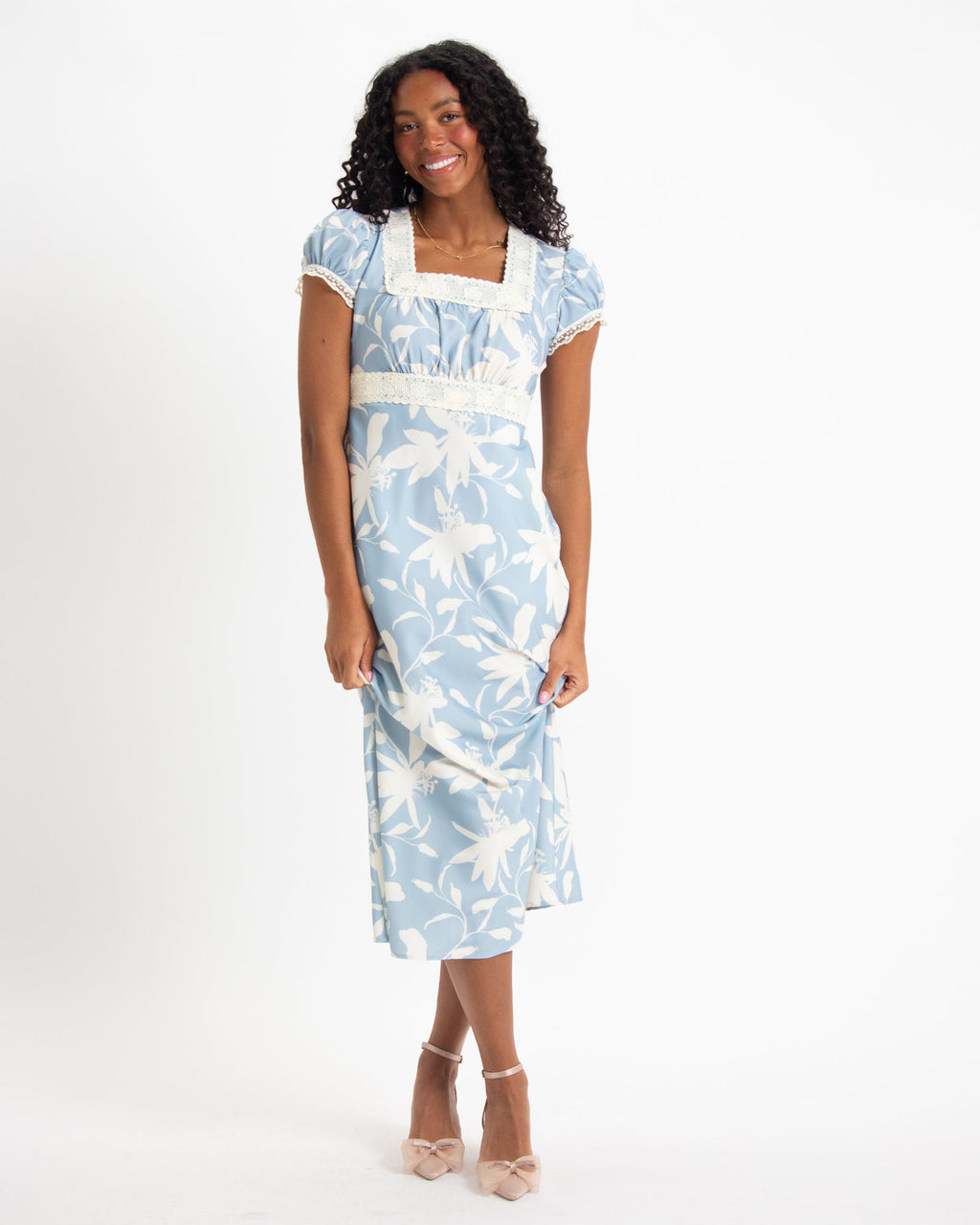 A woman wearing a light blue and white floral print midi dress with puff sleeves, square neckline, lace trim, and tie-back empire waist.