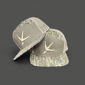 Side-by-side view of a 7-panel snapback cap featuring Mossy Oak New Bottomland camo, sage front panel, and embroidered turkey tracks logo.