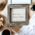 Rustic wooden sign with white background and black text that reads Wine: a hug in a glass, framed in dark distressed wood.