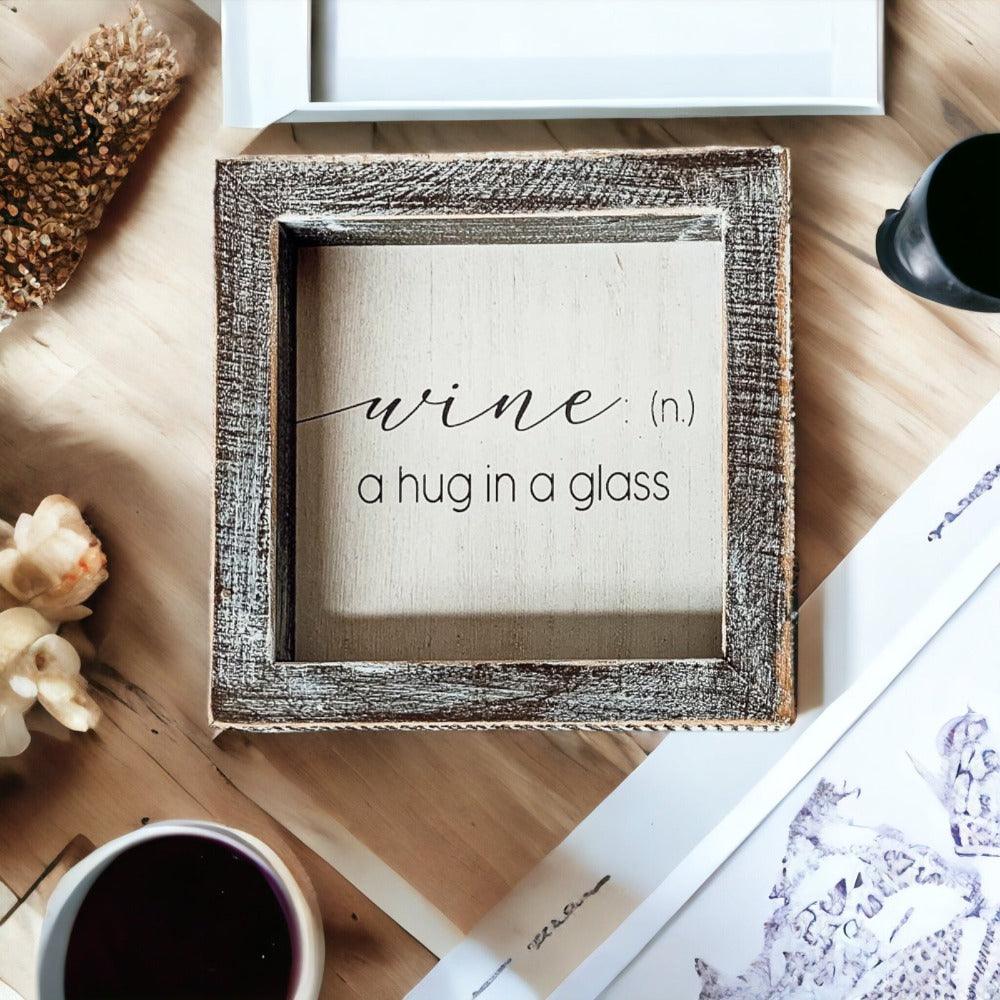 Rustic wooden sign with white background and black text that reads Wine: a hug in a glass, framed in dark distressed wood.