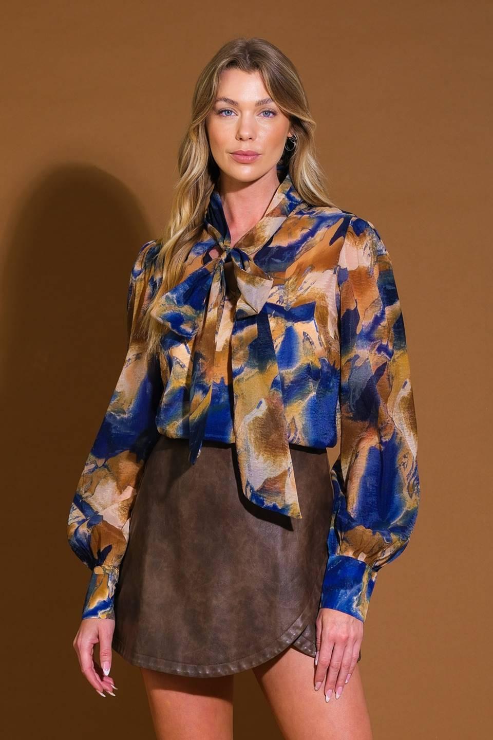 Model wearing a printed long-sleeve blouse with a tie-front neckline and blouson sleeves in blue and tan abstract print.