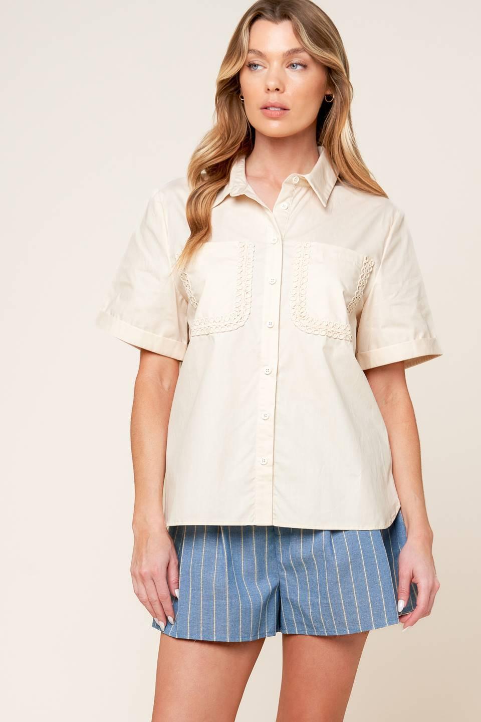 A woman wearing a cream button-down woven top with lace-trimmed pockets and cuffed short sleeves.