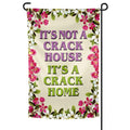 It's A Crack Home garden flag measuring 12 by 18 inches displayed on a metal stand in a green garden setting.