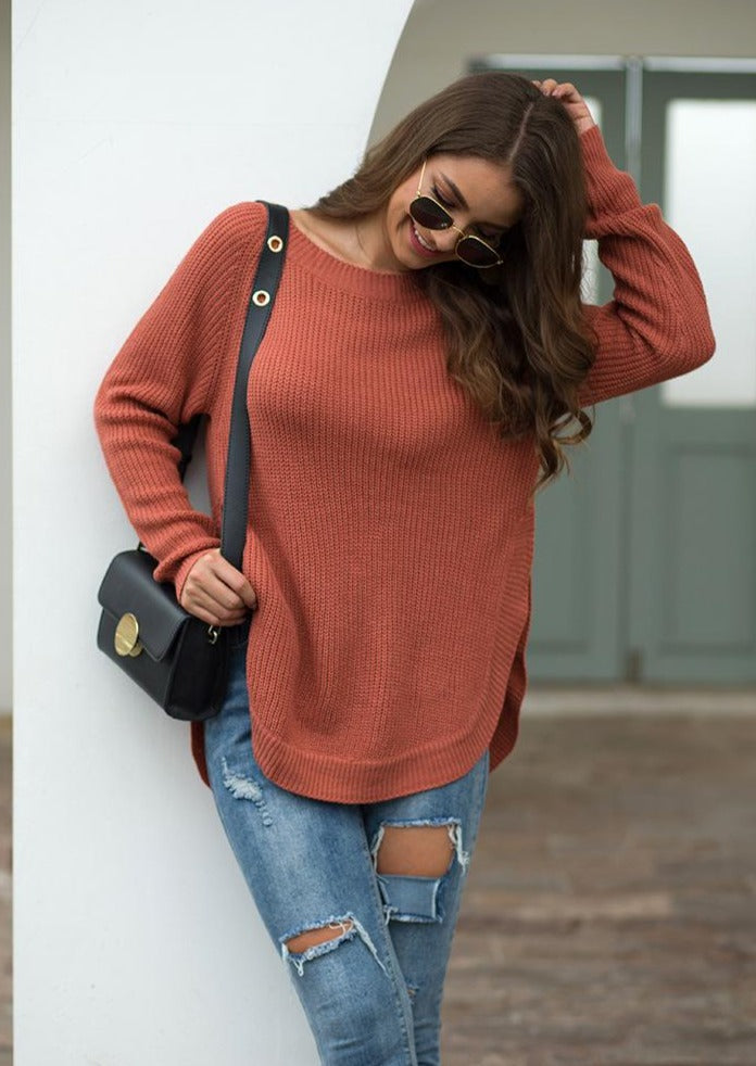 A woman wearing a rust-colored long sleeve chunky knit sweater with a rounded hem, paired with ripped jeans.