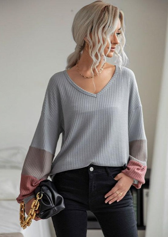 Model wearing a grey v-neck waffle knit sweater with colorblock striped sleeves styled with black jeans.
