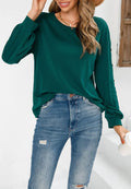 A woman wearing a long-sleeved dark green sweater with a round neck and side button details on the sleeves, styled with distressed blue jeans.