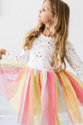 Young girl wearing a long sleeve dress with rainbow and unicorn print bodice and a multi-colored layered tulle tutu skirt.