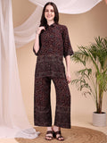 A woman wearing a maroon co-ord set featuring intricate floral and paisley motifs with a collar-neck shirt and palazzo pants.