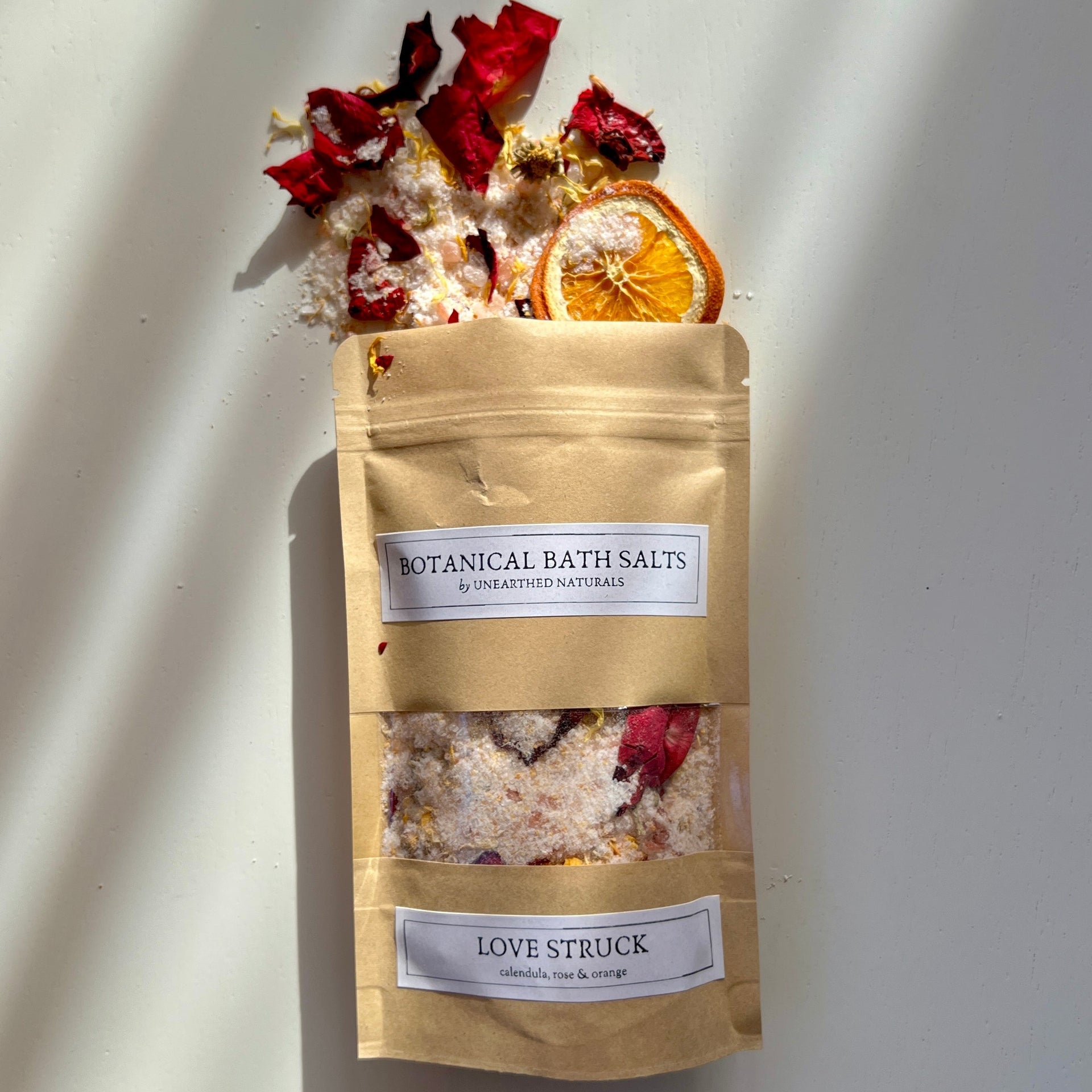 A 10 oz bag of Love Struck botanical bath salts surrounded by dried orange slices, rose petals, and calendula flowers on a wooden surface.