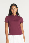 A woman wearing a deep fig-colored short-sleeve polo shirt with a mock collar and a pleated white skirt, shown from front and back views.