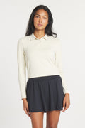A woman wearing a long-sleeve, Birch-colored Catalyst Polo with a point collar, paired with a black pleated skirt.