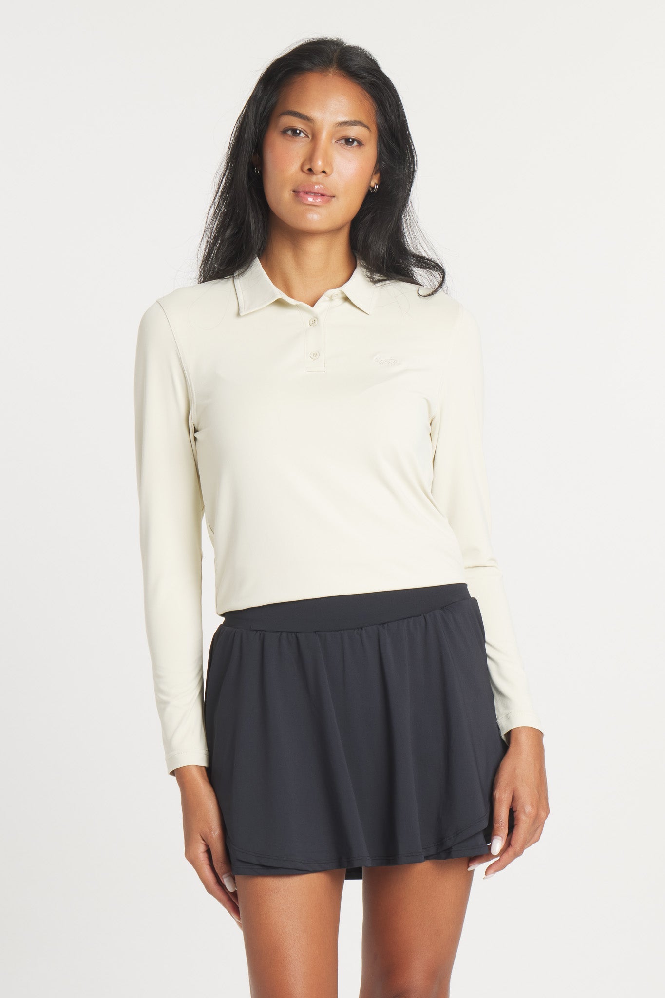 A woman wearing a long-sleeve, Birch-colored Catalyst Polo with a point collar, paired with a black pleated skirt.