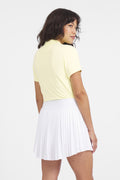 Women wearing a Buttercup yellow short-sleeve polo shirt with a mock collar, paired with a white pleated skirt.
