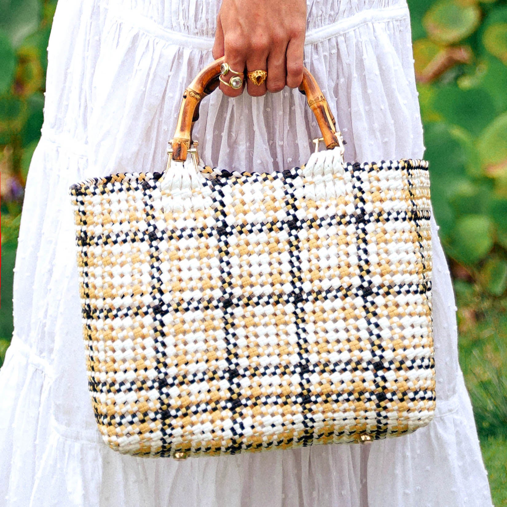 A woven recycled plastic market tote bag featuring a black, tan, and white plaid pattern with arched bamboo handles.